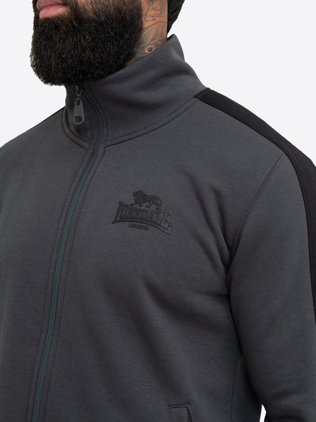 Nahaufnahme des anthrazitfarbenen Lonsdale Gummi-Logos auf der Brust der Trainingsjacke. 