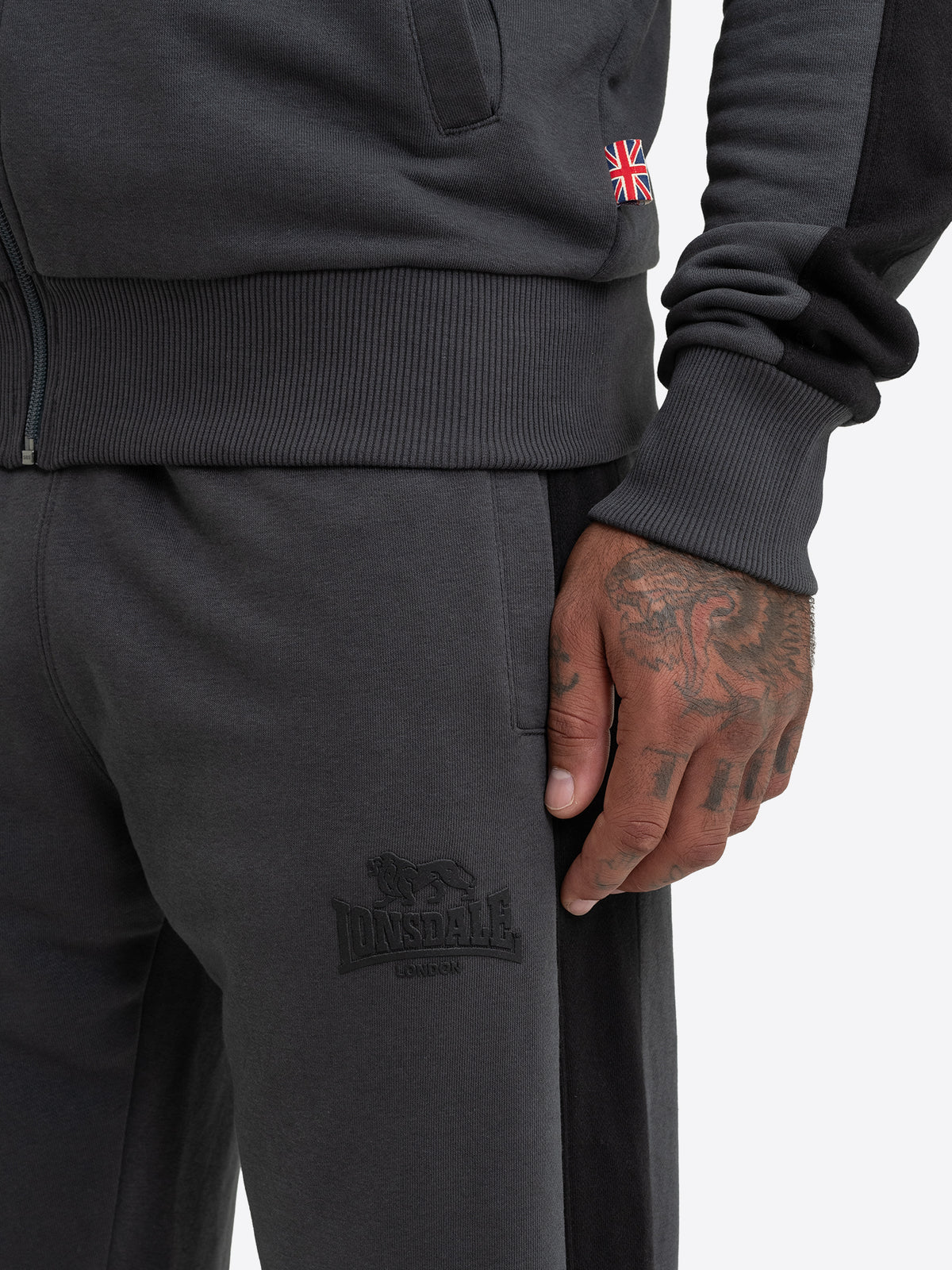 Nahaufnahme des anthrazitfarbenen Lonsdale Gummi-Logos auf dem Bein der Jogginghose