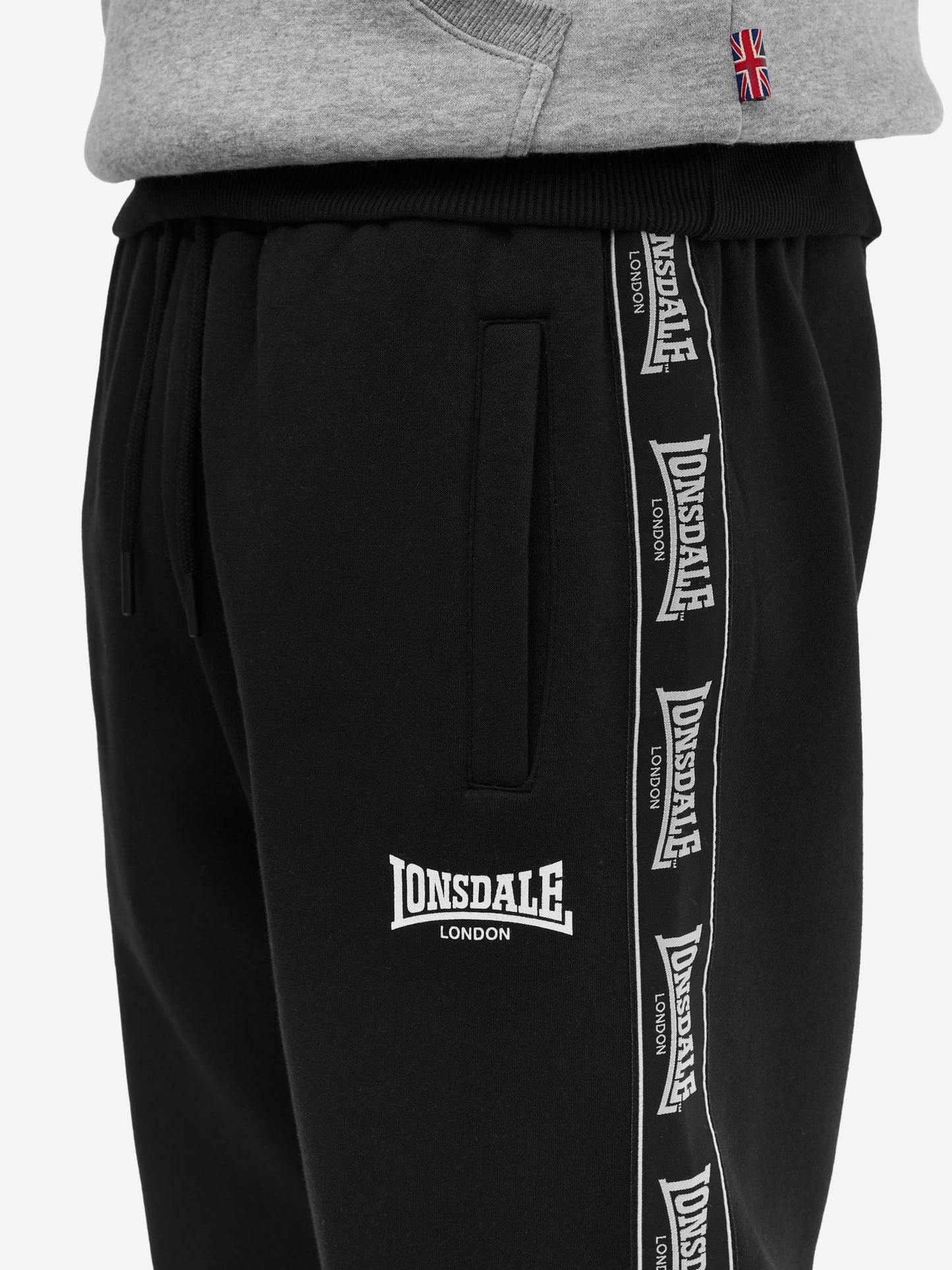 Nahaufnahme der Lonsdale Logo-Applikation und Fleece-Struktur des schwarz-grauen Jogginganzugs aus Baumwolle. 