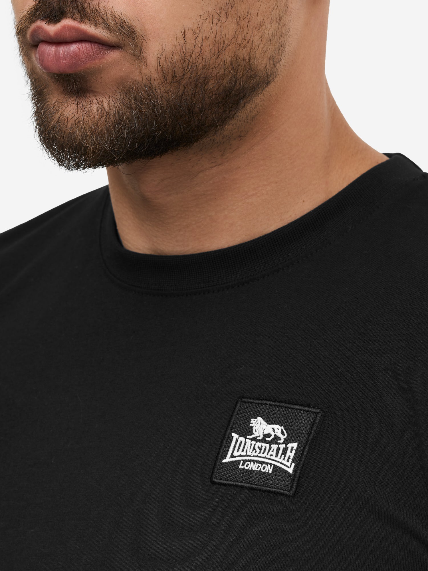 Nahaufnahme des Lonsdale-Patches auf schwarzer, gekämmter Baumwolle; zeigt das quadratische Logo-Label und die feine Jersey-Struktur.