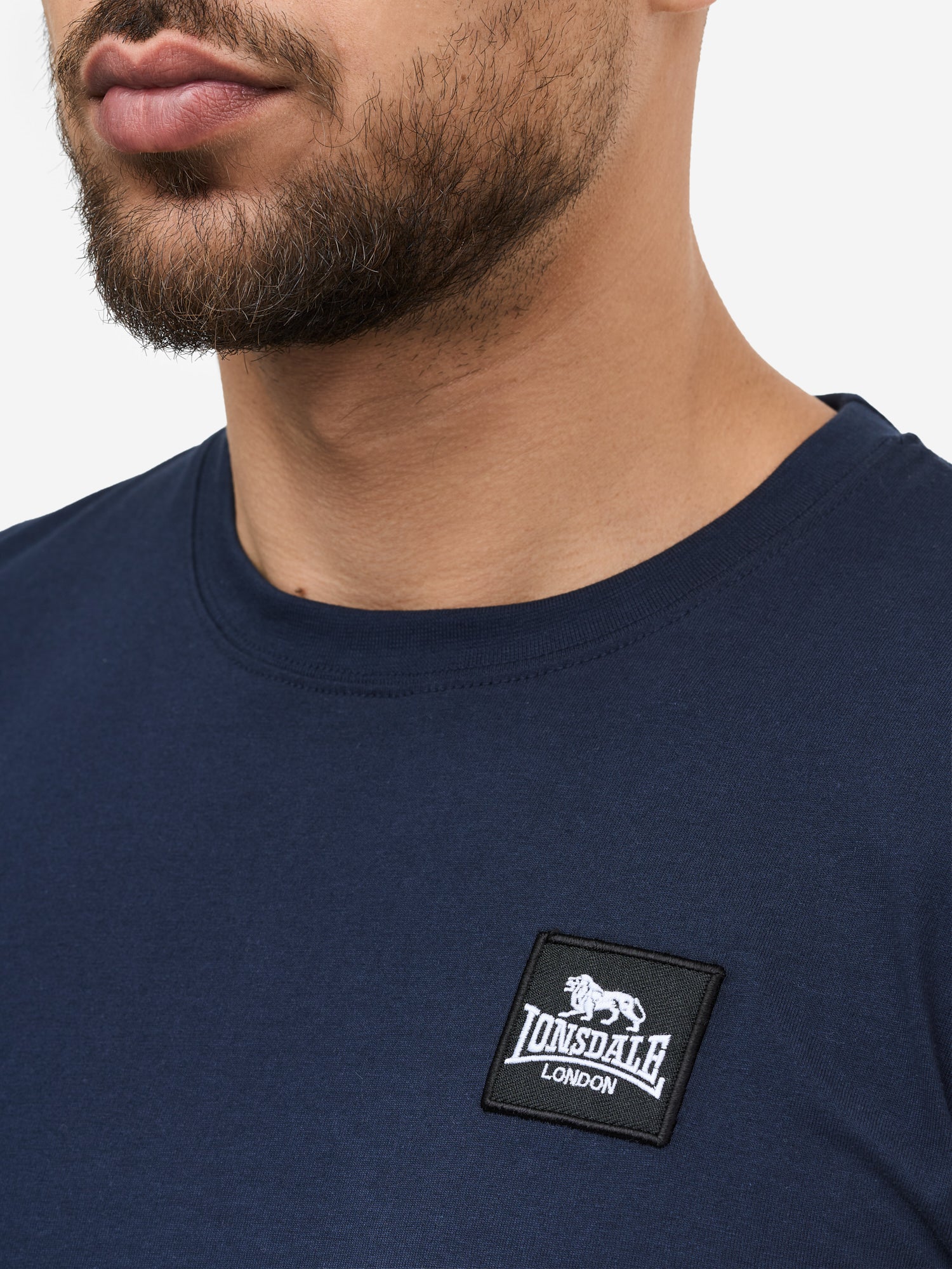 Nahaufnahme des Lonsdale-Patches auf dunkelblau, gekämmter Baumwolle; zeigt das quadratische Logo-Label und die feine Jersey-Struktur.