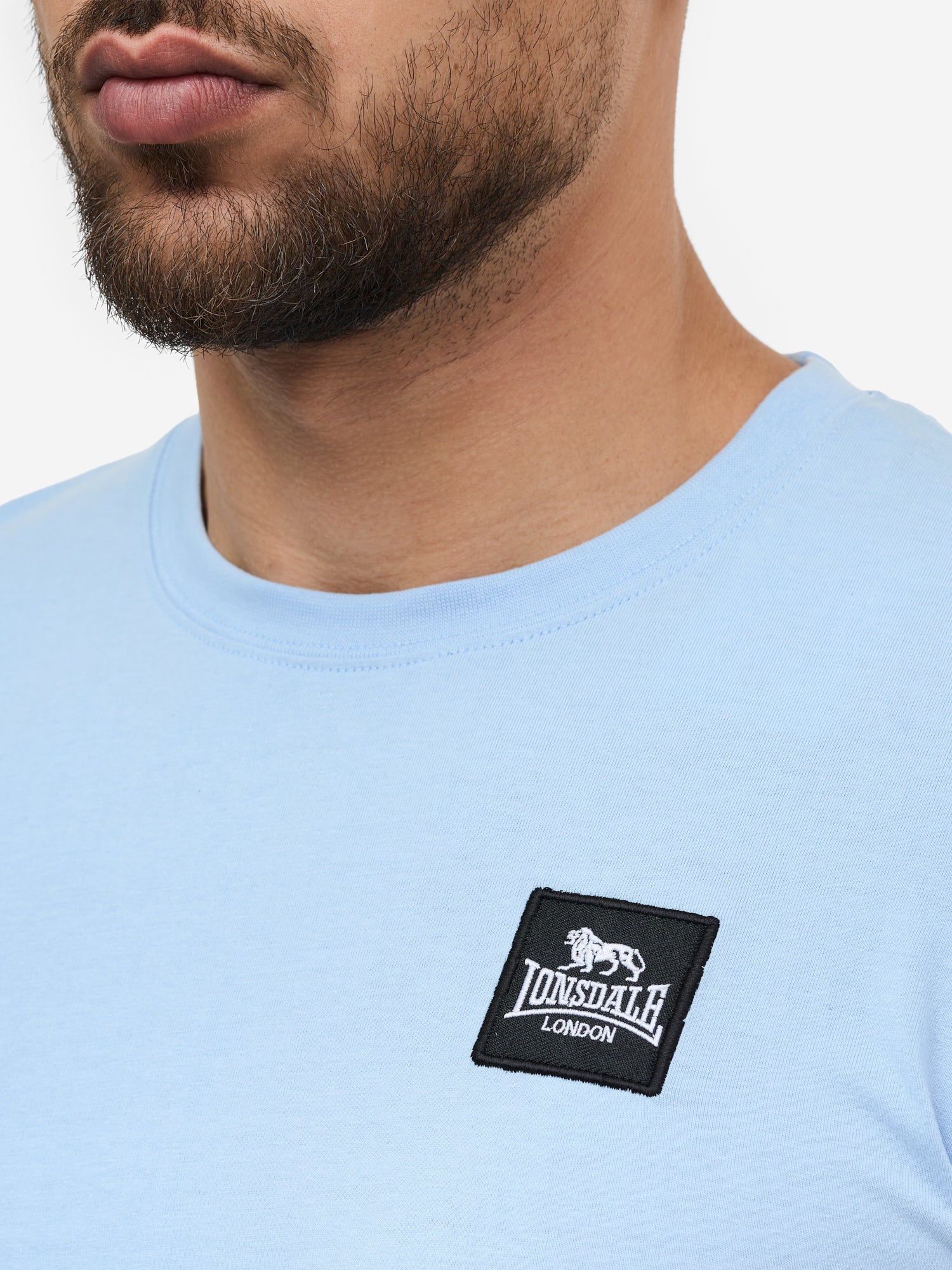 Nahaufnahme des Lonsdale-Patches auf blau, gekämmter Baumwolle; zeigt das quadratische Logo-Label und die feine Jersey-Struktur.