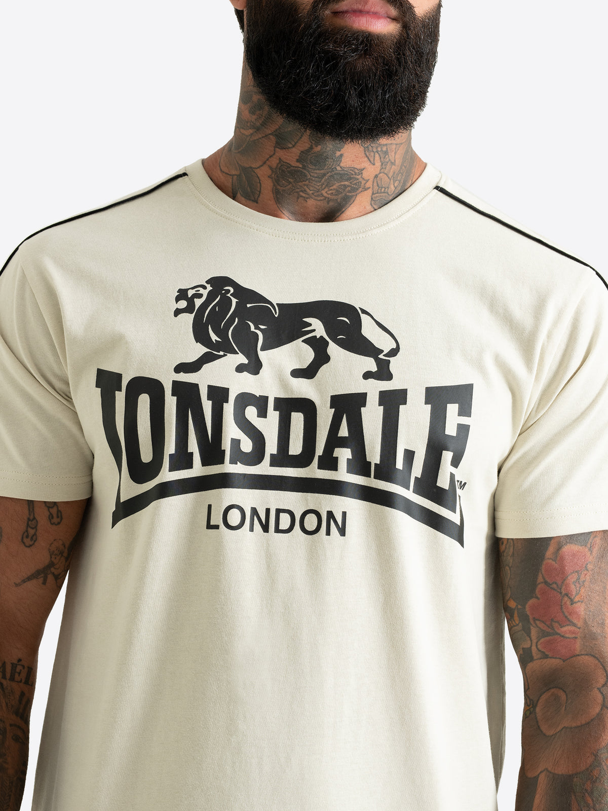 Nahaufnahme des schwarzen Lonsdale London Logo-Drucks auf der Brust des beigen Herren T-Shirts mit schwarzen Paspeln.