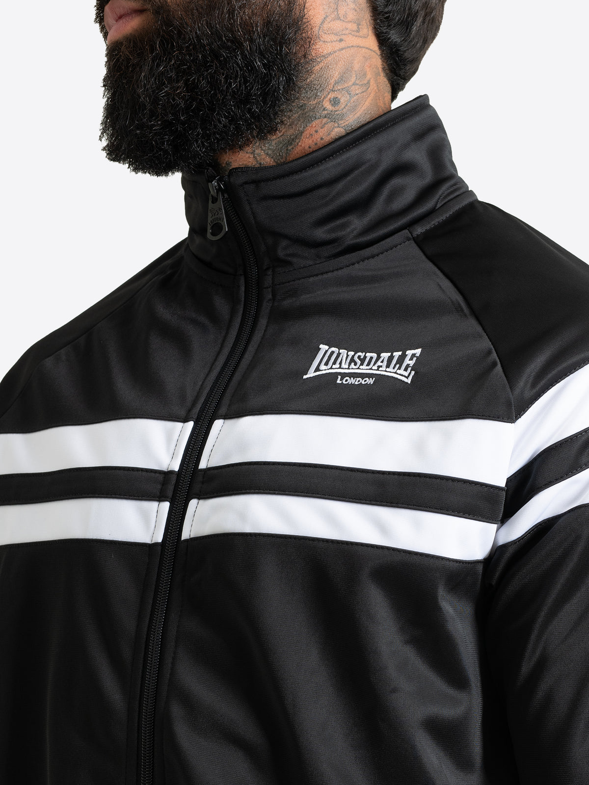 Detailansicht der schwarzen Lonsdale Herren-Trainingsjacke mit Fokus auf die weisse Logo-Stickerei und die Kontraststreifen.
