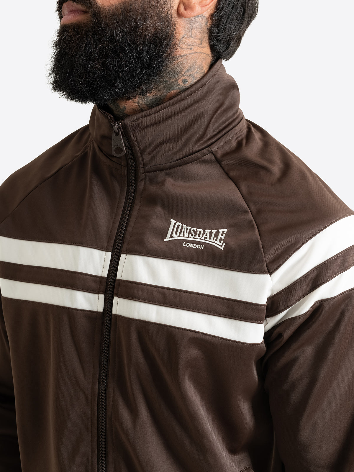 Detailansicht der braunen Lonsdale Herren-Trainingsjacke mit Fokus auf die weisse Logo-Stickerei und die Kontraststreifen.
