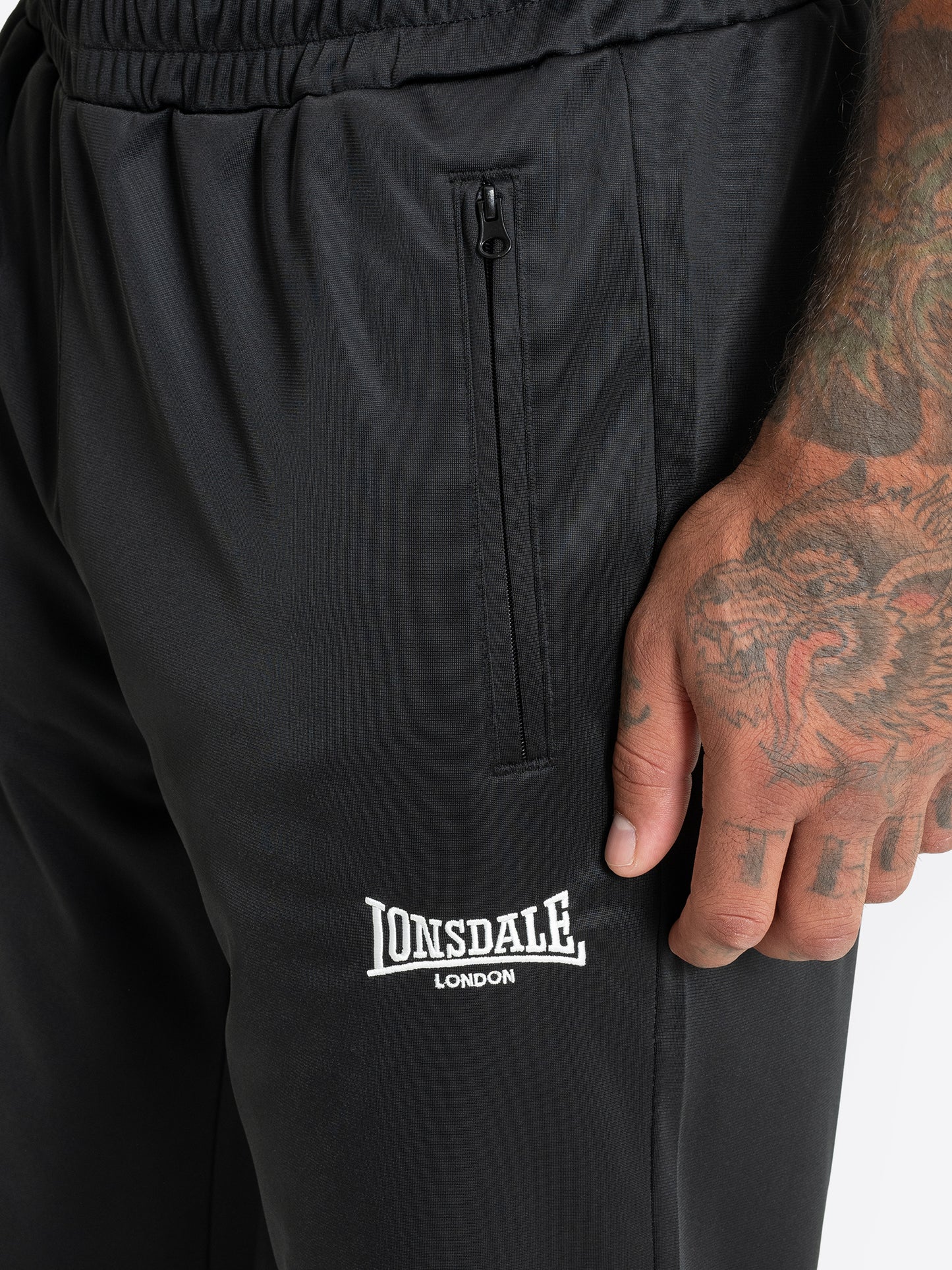 Detailansicht der Lonsdale Logo-Stickerei und Reissverschlusstasche der schwarzen Herren-Jogginghose.