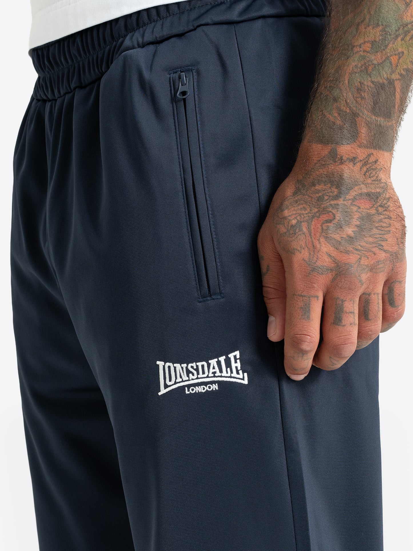Detailansicht der Lonsdale Logo-Stickerei und Reissverschlusstasche der dunkelblauen Herren-Jogginghose.