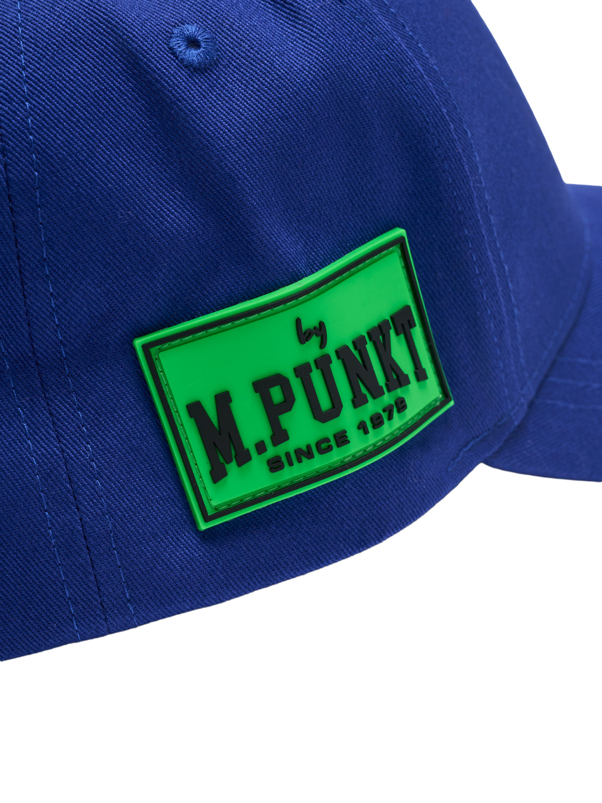 Nahaufnahme eines neongrünen Gummi-Patches mit der schwarzen Aufschrift „by M.PUNKT SINCE 1979“ auf einer blauen Kappe. 