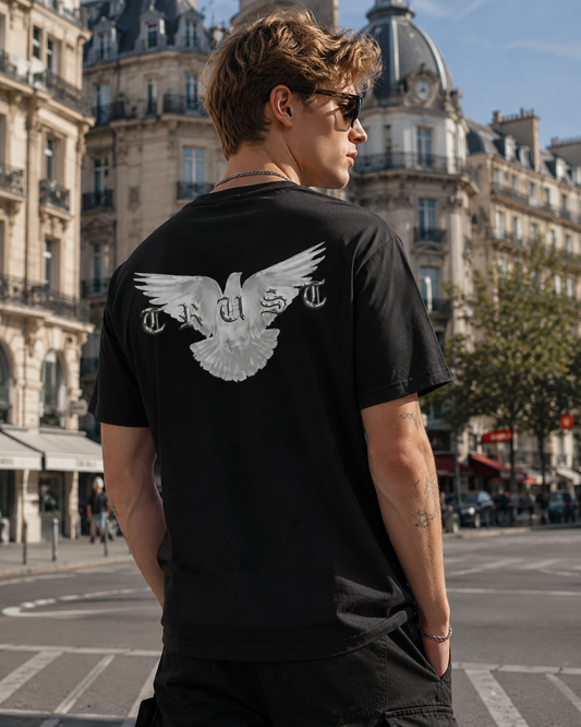 Junger blonder Mann in europäischer Altstadt trägt schwarzes Oversized Streetwear T-Shirt mit großem weißen Dove Backprint und TRUST Schriftzug im Urban Graphic Style.