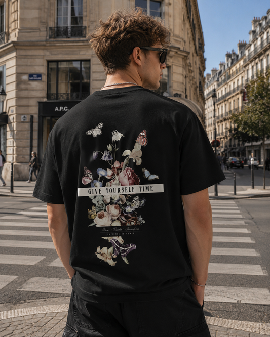 Junger Mann in europäischer Innenstadt trägt schwarzes Oversized Streetwear T-Shirt mit floralem Butterfly Backprint und „Give Yourself Time“ Schriftzug im modernen Urban Graphic Style.