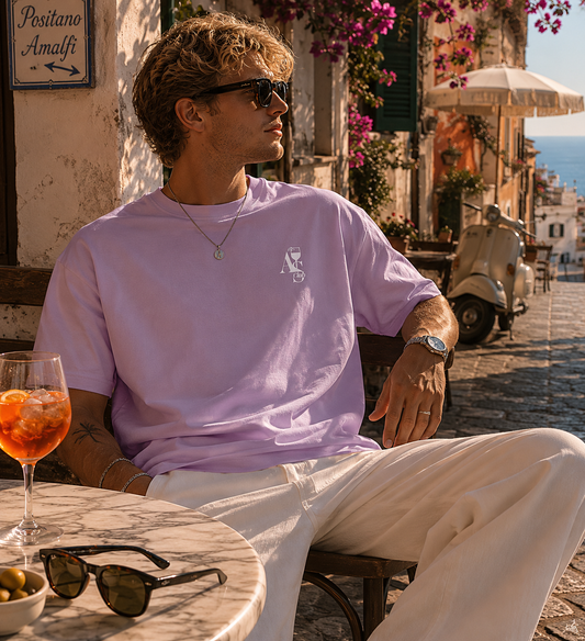Junger Mann trägt das lilacfarbene A S Club Tee mit kleinem minimalistischen Frontprint in einer sonnigen italienischen Küstenstadt. Sommerlicher Aperitivo Lifestyle mit mediterraner Café-Atmosphäre, Spritz Drink und entspanntem Vacation Look.