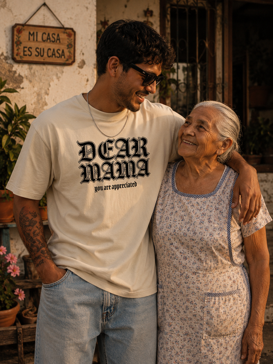 Junger Mann trägt das Dear Mama Tee in Sand mit schwarzem Frontprint neben einer älteren Frau vor einem mediterranen Haus bei goldenem Sonnenuntergang.